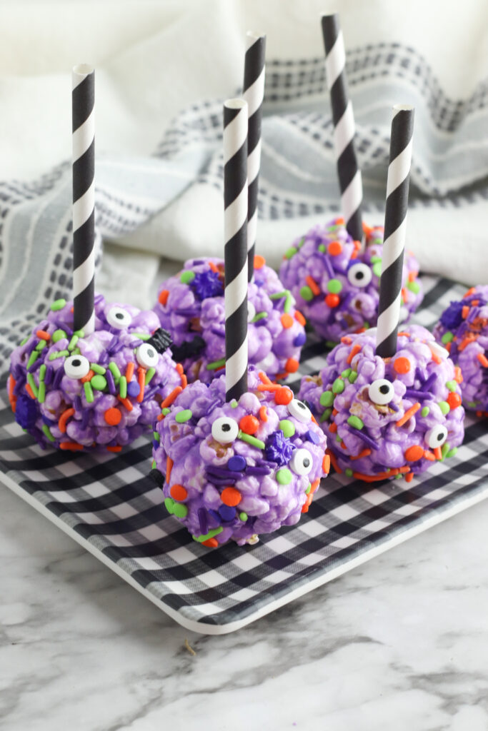 6 Creepy Halloween Popcorn Balls - purple colored popcorn balls with candy Halloween sprinkles and candy eyes - on a buffalo plaid plate with black and white straws to serve.