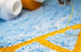 A machine embroiders the letter "A" in gold thread on light blue fabric. More thread spools are in the background.