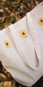 A white canvas tote bag is positioned upright on a pile of fall leaves. The bag has three embroidered sunflowers.