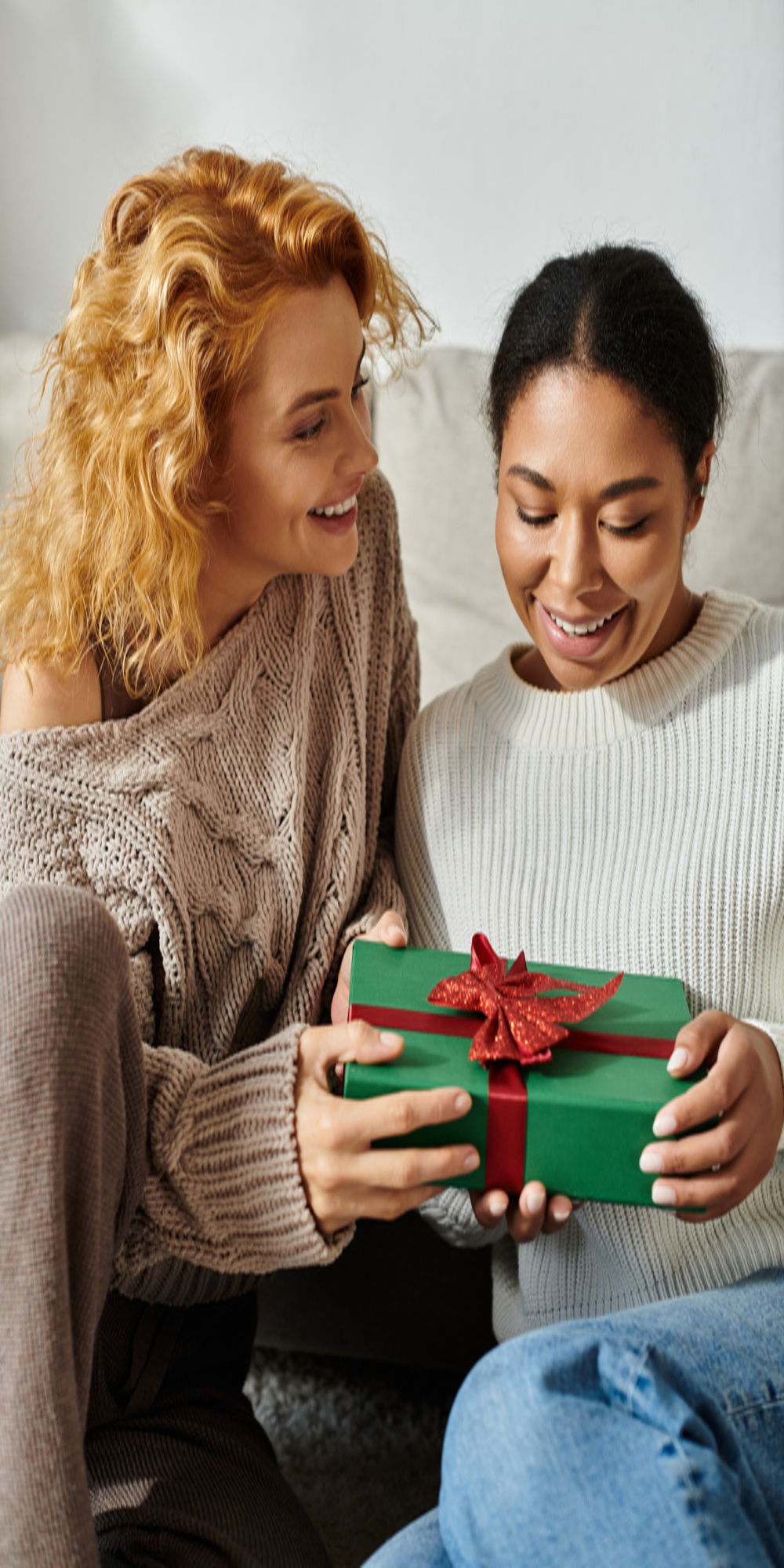 An adult woman smiles as she hands a gift wrapped in green paper with a red bow to a smiling teenage girl.
