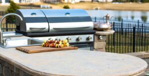 A close-up of a rock countertop with a tray full of vegetable skewers, next to a stainless-steel sink and a grill.