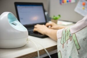 A close-up view shows a white and teal breast pump motor sitting on the desk of a woman who's typing on a laptop.