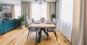 A dining room with wooden floors, five fabric chairs, placement mats, a bowl with fruit, and large white curtains.
