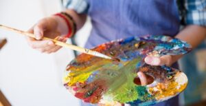 A woman in a smock holding an artist's palette. The palette is smeared with a wide variety of paint colors.