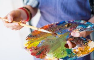 A woman in a smock holding an artist's palette. The palette is smeared with a wide variety of paint colors.