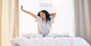 A woman is sitting up in bed and stretching out her arms. Her eyes are closed and there is a smile on her face.