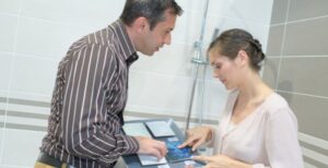 A man and woman standing in a bathroom shower stall while looking at samples of ceramic tile materials.
