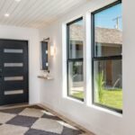 An entry way with a brown door, an area rug, two vertical windows, a small sitting area to the right and wall lights.