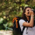 A woman with a black and orange backpack looking through the viewfinder of her camera to take a picture.