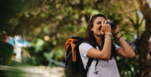 A woman with a black and orange backpack looking through the viewfinder of her camera to take a picture.
