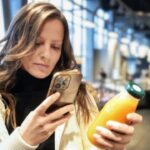 A woman stands in a supermarket holding a bottle of orange juice in one hand and her smartphone in the other.
