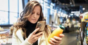 A woman stands in a supermarket holding a bottle of orange juice in one hand and her smartphone in the other.