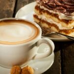 A cup of coffee in a white mug on a white saucer sits next to a layered chocolate cake dessert. There's a fork with the cake.