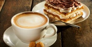 A cup of coffee in a white mug on a white saucer sits next to a layered chocolate cake dessert. There's a fork with the cake.