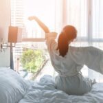 A woman stretching while sitting on a white bed in a bright white room with pillows, sunlight, and curtains.