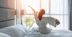 A woman stretching while sitting on a white bed in a bright white room with pillows, sunlight, and curtains.