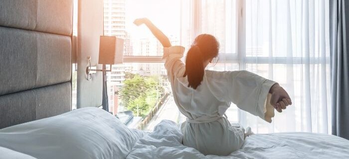 A woman stretching while sitting on a white bed in a bright white room with pillows, sunlight, and curtains.
