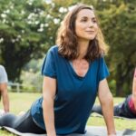 A woman smiles while practicing yoga outdoors in a park as part of a class. Three other people perform yoga around her.