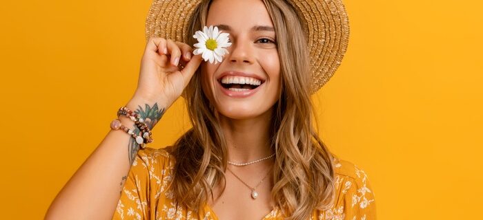 A smiling woman in a straw hat holds a white daisy over one eye. She wears a yellow floral dress and layered necklaces.